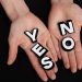 Two hands holding black and white letters spelling 'YES' and 'NO' on a black background.